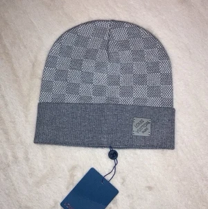 Grå mössa med rutmönster - Snygg grå mössa med klassiskt rutmönster och uppvikt kant. Mössan har en diskret logotyp på framsidan och är tillverkad i mjukt material som känns lyxigt. Perfekt accessoar för att lyfta din stil.