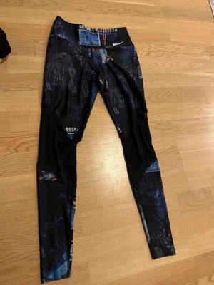 Nike leggings med grafiskt mönster - Säljer ett par Nike leggings med coolt grafiskt mönster i svart, blått och inslag av rött och vitt. De har hög midja och är tillverkade i stretchigt material som passar perfekt för träning eller chill. Nike-logga framtill ger extra sportig vibe.