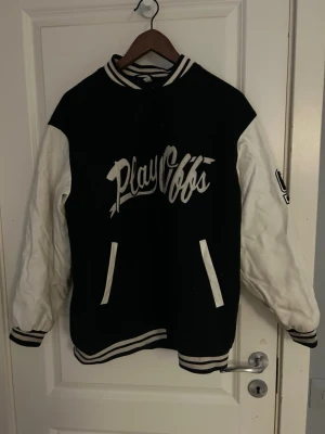 Svartvit bomberjacka  - Säljer en svartvit bomberjacka från Divided i storlek S. Jackan har vita ärmar, svart kropp och broderad text 'Play Offs' samt siffran 92 på ena ärmen. Klassiska ränder på muddar och krage, stängs med knappar framtill. Perfekt för en sportig och trendig look.