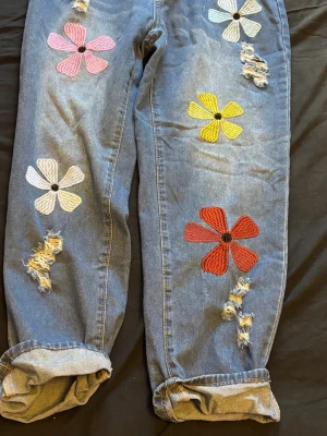 Blommiga boyfriend jeans med broderi - Unika blå jeans med färgglada broderade blommor i rött, gult, vitt och rosa. Modellen är loose fit med uppvikta benslut och slitna detaljer på framsidan. Perfekta för dig som vill sticka ut med en kreativ och lekfull stil.