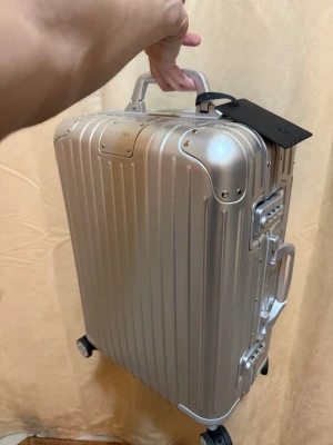 Silver resväska från Rimowa - Snygg och stilren silverfärgad resväska från Rimowa i aluminium. Väskan har fyra hjul, flera handtag och klassisk räfflad design. Perfekt för dig som vill resa med stil och ha en tålig kabinväska. Märkets logga syns på väskan och det finns en svart bagagetagg.