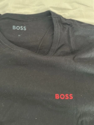 Svart BOSS t-shirt med röd logga - Säljer en svart t-shirt från BOSS med klassisk rund hals och korta ärmar. T-shirten har en stilren röd BOSS-logga broderad på bröstet. Tillverkad i mjuk bomull för skön känsla och enkel att matcha med allt.