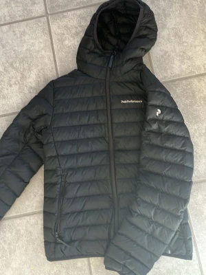 Svart dunjacka Peak Performance med huva - Snygg svart dunjacka från Peak Performance med huva och dragkedja framtill. Jackan har två sidofickor med dragkedja och diskret logga på bröstet samt ärmen. Perfekt för kalla vinterdagar tack vare det värmande dunmaterialet och den stilrena designen.