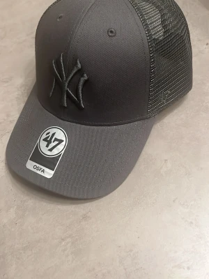 Mörkgrå Yankees keps från '47 Brand - Snygg mörkgrå keps med New York Yankees-logga i svart brodyr framtill. Kepsen har böjd skärm och mesh-paneler på sidorna och bak för extra ventilation. Justerbar snapback baktill så den passar de flesta. Perfekt för en sportig och avslappnad stil. Helt oanvänd köpt på zalando för 1mån sedan.