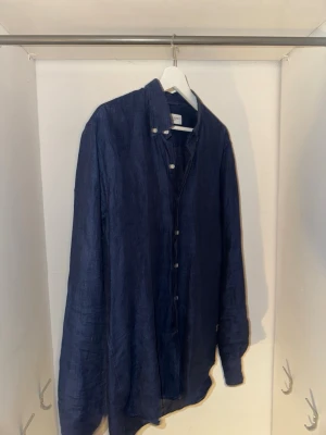 John Henric linneskjorta - Snygg mörkblå skjorta i luftigt linne med klassisk krage och vita knappar hela vägen framtill. Skjortan har lång ärm och en avslappnad passform, perfekt för en stilren och bekväm look.