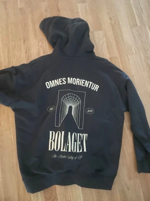 Svart hoodie från Bolaget - Svart hoodie med stor vit print på ryggen med texten 'OMNES MORIENTUR' och 'BOLAGET'. Snygg grafisk design och huva. Perfekt för dig som gillar streetwear och vill sticka ut med en unik look.