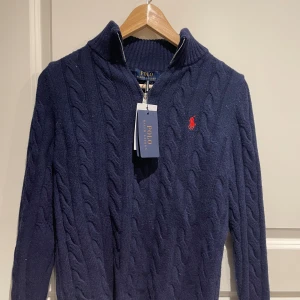 Mörkblå kabelstickad tröja Polo Ralph Lauren - Mörkblå kabelstickad tröja från Polo Ralph Lauren med halv dragkedja och klassisk röd logga broderad på bröstet. Tröjan har hög krage och ribbade muddar. Perfekt för lager på lager och en stilren look.