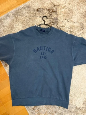  Nautica tröja  - Blå sweatshirt från Nautica med tryck 'Nautica EST. 1983' på bröstet. Tröjan har en loose passform, rund halsringning och långa ärmar. Perfekt för en avslappnad stil.