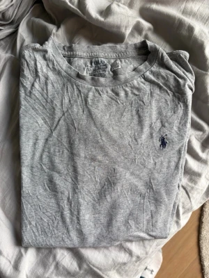 Grå t-shirt från Polo Ralph Lauren - Klassisk grå t-shirt från Polo Ralph Lauren med rund hals och den ikoniska lilla broderade loggan på bröstet. Tillverkad i mjuk bomull och har en enkel, clean look som passar till allt. Perfekt basplagg för garderoben.Strl passar Small även XS den har dock ett litet litet hål i t shirten
