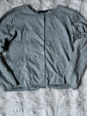 Grå basic kofta med knappar - En enkel och stilren grå kofta med rund halsringning och knäppning framtill. Långärmad modell i mjukt material, perfekt att slänga över en t-shirt eller linne. Klassisk passform och ribbade muddar vid ärmslut och nederkant.