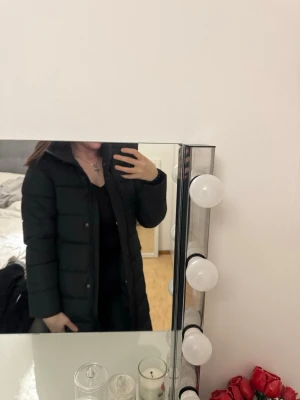 Jacka - Säljer en svart lång dunjacka med huva och quiltad design. Jackan har dragkedja framtill och stora fickor på sidorna. Perfekt för kalla vinterdagar och ger en stilren look.