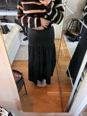 Svart långkjol med volangkant - Svart långkjol med bred midjeresår och volangkant nertill. Kjolen har en flowy passform och är perfekt för dig som gillar en enkel men snygg stil. Tillverkad i ett mjukt och lätt material som känns skönt mot huden.