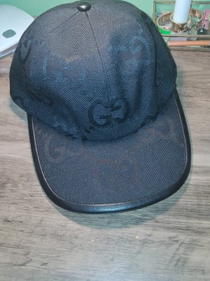 Svart keps från Gucci med GG-mönster - Snygg svart keps från Gucci med klassiskt GG-mönster över hela kepsen. Kepsen har böjd skärm med svart kant i läderimitation och är tillverkad i ett tåligt tygmaterial. Perfekt accessoar för att lyfta din streetwear-look. DM f