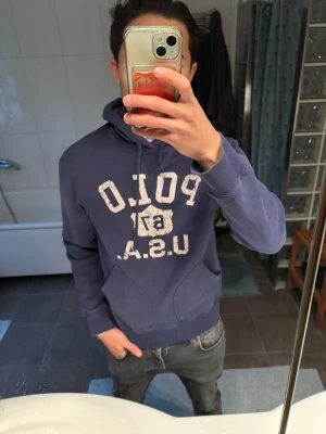Blå hoodie från Polo USA - Säljer en blå hoodie från Polo Ralph Lauren USA med vit, sliten logotyp på bröstet och klassisk huva med dragsko. Hoodien har en stor magficka och ribbade muddar vid ärmslut och nederkant. Perfekt för en avslappnad och cool look.