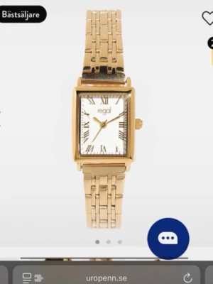 Guldfärgad klocka från Regal - Stilren guldfärgad klocka från Regal med fyrkantig urtavla och romerska siffror. Klockan har en klassisk länkarmband i metall och vit urtavla som ger en elegant look. Perfekt accessoar för att lyfta din outfit.