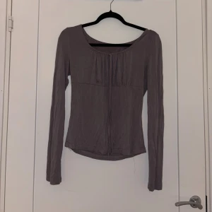 Ljuslila/grå långärmad topp - Säljer en ljuslila långärmad topp med rundad halsringning och fina veckdetaljer framtill. Modellen är figurnära och har en mjuk, tunn känsla som passar perfekt till jeans eller kjol.