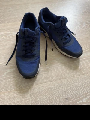 Mörkblå Nike sneakers - Snygga mörkblå Nike sneakers med lågt skaft och vita sulor. Skorna har klassisk snörning, diskret Nike-logga på sidan och ovandel i mesh och syntetmaterial. Perfekta för dig som gillar sportig stil och vill ha bekväma skor till vardags. Storlek 44