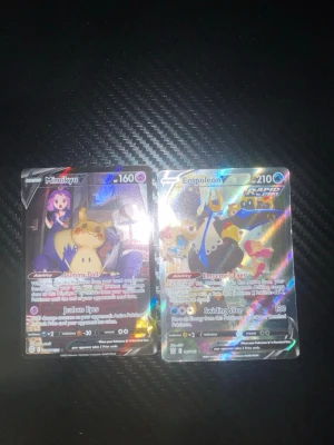 Pokémon holografiska samlarkort - Två holografiska Pokémon samlarkort: Mimikyu V och Empoleon V. Kortens framsida har färgstarka motiv och glansig finish, baksidan är blå med Pokéball-design. Perfekt för samlare eller trading card game-entusiaster.