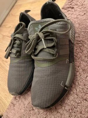 Adidas NMD R1 olivgröna sneakers - Säljer ett par Adidas NMD R1 sneakers i olivgrönt tyg med matchande snören och detaljer. Skorna har en modern, sportig look med textilöverdel och klassiska NMD-pluggar på sulan. Perfekta för dig som gillar streetwear och vill ha något bekvämt och stilrent.