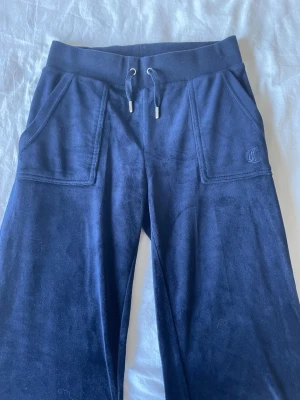 Mörkblå mjukisbyxor från Juicy Couture - Mörkblå mjukisbyxor i velour från Juicy Couture med resår i midjan och dragsko. Fickor fram och bakfickor med knappar. Liten JC-logga broderad på ena fickan fram. Perfekta för en chill och avslappnad stil.