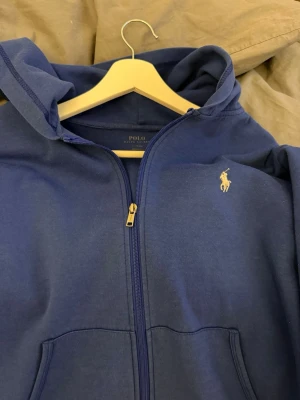 Blå hoodie från Polo Ralph Lauren - Snygg blå hoodie från Polo Ralph Lauren med dragkedja framtill och klassisk huva. Ikoniska loggan i vitt broderad på bröstet. Hoodien har kängurufickor och är gjord i mjukt bomullsmaterial. Perfekt för en avslappnad och stilren look.