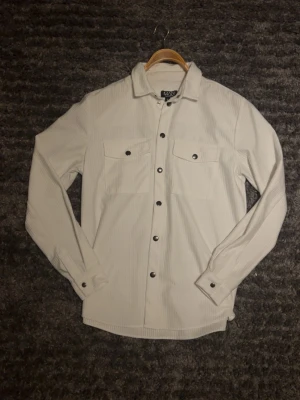 Vit overshirt från MAN - Stilren vit overshirt från MAN med diskreta ränder, två bröstfickor sopknappar och klassisk kr sopknappar och klassisk krage. Långärmad modell i ett mjukt tyg som passar perfekt för lager-på-lager. Snygga svarta knappar ger en modern touch.