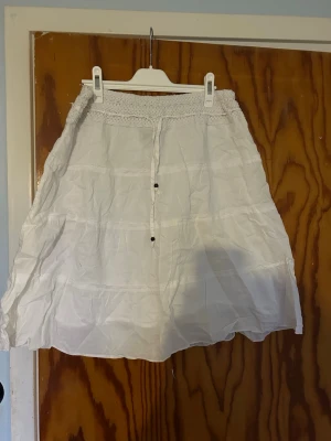 Vit långkjol med spetsdetalj - Luftig vit långkjol med bred resår i midjan och spetsdetalj upptill. Kjolen har snören framtill och är sydd i flera våder för en bohemisk look. Perfekt för varma dagar och enkel att matcha med olika toppar.