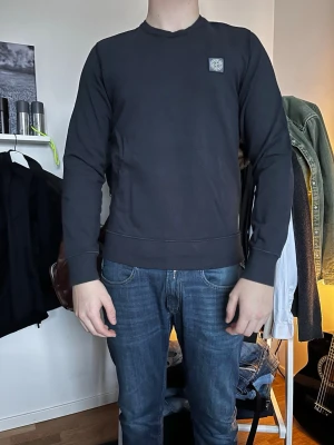 Svart sweatshirt från Stone Island - Säljer en svart sweatshirt från Stone Island med rund hals och diskret logopatch på bröstet. Tröjan har långa ärmar, ribbade muddar och är gjord i mjukt bomullsmaterial. Perfekt för en clean och stilren look.