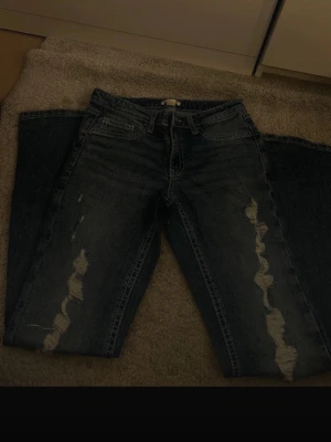 Mörkblå slitna jeans med raka ben - Säljer ett par mörkblå jeans med raka ben och slitningar längs framsidan. Jeansen har klassisk femficksdesign och tydliga kontrastsömmar. Perfekta för en avslappnad och trendig look.