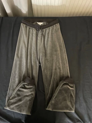 Grå velourmjukisbyxor med muddar - Mjukisbyxor i grå velour med breda muddar i mörkare grått vid midja och benslut. Byxorna har en avslappnad passform och är perfekta för att chilla hemma. Materialet är mjukt och skönt mot huden.