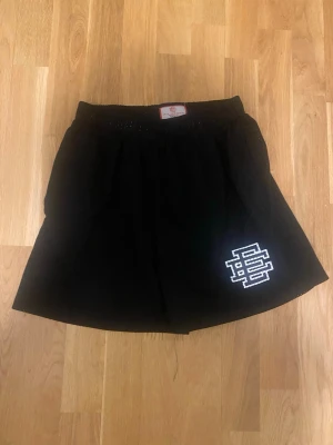 Svarta meshshorts från Eric Emanuel - Svarta shorts från Eric Emanuel i meshmaterial med elastisk midja och vit logga på ena benet. Perfekta för en sportig look och riktigt sköna att bära. Klassisk design med luftig känsla.