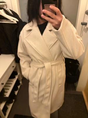 Ljusbeige kappa med skärp - Stilren ljusbeige kappa med breda slag och knytskärp i midjan. Kappan har rak passform, dolda fickor och är tillverkad i ett mjukt tyg som ger en lyxig känsla. Perfekt för dig som gillar minimalistisk och trendig stil. Passar vår och höst perfekt! 