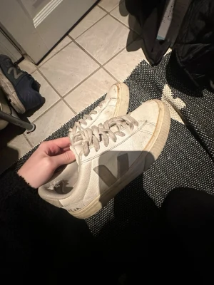Vita Veja sneakers med grå detalj - Säljer ett par klassiska vita Veja sneakers med grå V-logga på sidan. Skorna har låga skaft, vita snören och sula i naturfärgad gummi. Tillverkade i läder med textildetaljer. Perfekta för dig som gillar stilrena och hållbara sneakers. 
