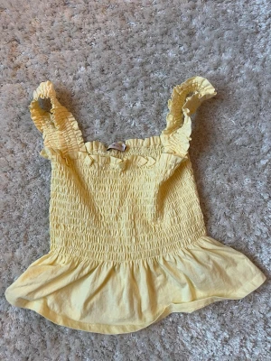 Gul smockad linnetopp med volang - Söt gul linnetopp med smockad överdel och breda axelband. Toppen har volangkant nedtill och rynkade detaljer vid axelbanden. Perfekt för soliga dagar och ger en fräsch look.