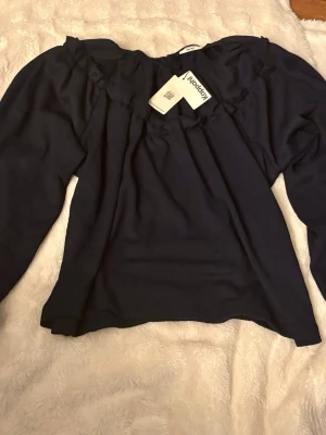 Mörkblå offshoulder blus KappAhl - Trendig mörkblå offshoulder blus från KappAhl med långa ärmar och volangdetaljer längs ringningen. Blusen har en lös passform och är tillverkad i ett mjukt, lätt material som ger en skön känsla. Perfekt att styla med jeans eller kjol.