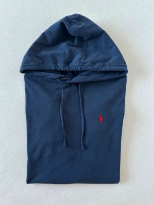Mörkblå hoodie från Polo Ralph Lauren -  Pris kan diskuteras. Snygg mörkblå hoodie från Polo Ralph Lauren med klassisk röd logga broderad på bröstet. Hoodien har huva med dragsko och långa ärmar. Tillverkad i mjukt bomullsmaterial som känns skönt mot huden. Perfekt för en avslappnad och stilren look. Den har dock ett litet hål på ärmen. 