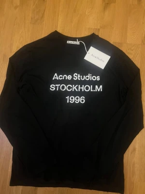 Svart långärmad topp Acne Studios - Svart långärmad topp från Acne Studios med vit text 'Acne Studios STOCKHOLM 1996' på bröstet. Klassisk rund hals och relaxed fit. Perfekt för dig som gillar stilrena och trendiga plagg med tydlig logga.
