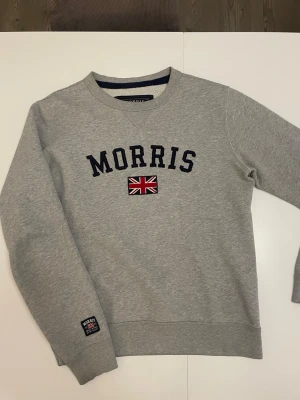Morris tröja  - Snygg grå sweatshirt från Morris med broderad logga och brittisk flagga på bröstet. Tröjan har rund halsringning, ribbade muddar och en patch på ärmen. Perfekt för en avslappnad och stilren look.