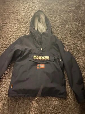 Mörkblå vindjacka från Napapijri - Snygg mörkblå vindjacka från Napapijri med halv dragkedja, stor huva och framficka med norsk flagga. Jackan har logotyp på bröstet och patch på ärmen. Materialet är tåligt och vattenavvisande, perfekt för blåsiga dagar.