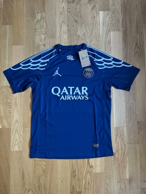 Blå PSG x Jordan matchtröja - Snygg blå PSG matchtröja i samarbete med Jordan. Tröjan har vita detaljer, vingmönster på axlarna och Qatar Airways-logga på bröstet. Tillverkad i lätt och ventilerande funktionsmaterial, perfekt för fotboll eller träning.