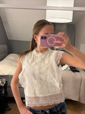 Vit spetsblus med volangdetaljer - Superfin vit blus i spets med volangdetaljer vid ärmslut och nederkant. Blusen är ärmlös och har en hög hals med dekorativ kant. Perfekt att styla med jeans eller kjol för en trendig look.                   Storleken är S men skulle säga att den sitter mer som Xs