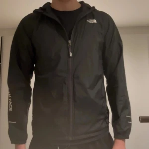 Svart vindjacka från The North Face - Svart vindjacka från The North Face med huva och dragkedja framtill. Jackan har logga på bröstet och ärmen, samt reflekterande detaljer på ärmarna. Tillverkad i lätt polyester, perfekt för blåsiga dagar. Snygg och enkel design som funkar till det mesta.