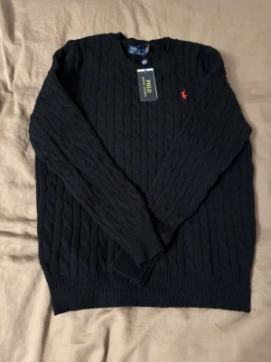 Svart kabelstickad tröja Polo Ralph Lauren - Kabelstickad svart tröja från Polo Ralph Lauren med rund halsringning och röd broderad logga på bröstet. Tröjan har långa ärmar och ribbade muddar vid ärmslut och nederkant. Perfekt för dig som gillar klassisk och stilren look.