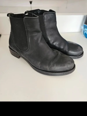 Svarta Chelsea boots i skinn - Klassiska svarta Chelsea boots i äkta skinn med elastiska paneler på sidorna och låg klack. Tidlös design som passar till det mesta och ger en clean look. Perfekta för dig som gillar enkel och stilren stil. Se bilder för Skavanker. Läder. 