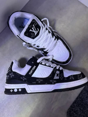 Louis Vuitton svartvita sneakers - Snygga sneakers från Louis Vuitton i svart och vitt med klassiskt LV-mönster. Skorna har vita snören, svartvit sula och tydliga logodetaljer på plös och sidor. Materialet är en mix av läder och textil för en exklusiv känsla.