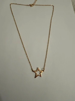 Guldfärgat halsband med stjärnhänge - Snyggt halsband i guldfärg med en tunn kedja och ett öppet stjärnformat hänge. Perfekt accessoar för att ge din outfit en trendig touch. Halsbandet har en enkel och stilren design som passar till många olika looks.