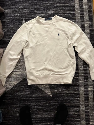 Vit sweatshirt från Polo Ralph Lauren - Klassisk vit sweatshirt från Polo Ralph Lauren med rund halsringning och den ikoniska lilla broderade loggan på bröstet. Tröjan har långa ärmar och ribbade muddar vid ärmslut och nederkant. Perfekt för en clean och stilren look.