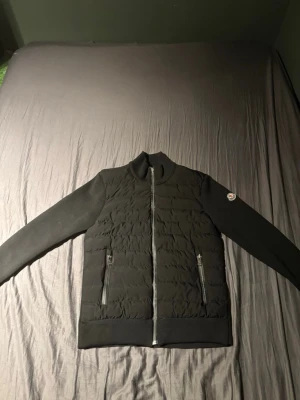 Svart pufferjacka från Moncler - Svart pufferjacka från Moncler med quiltad front och släta ärmar. Jackan har två dragkedjeförsedda fickor och en hög krage. Moncler-logga på ena ärmen. Perfekt för dig som vill ha en stilren och sportig look.