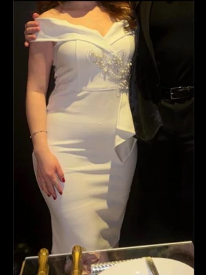 Vit kortklänning med pärldetaljer - Elegant vit kortklänning med offshoulder-design och figurnära passform. Klänningen har dekorativa pärlor och volangdetalj framtill. Passer för både M och L 