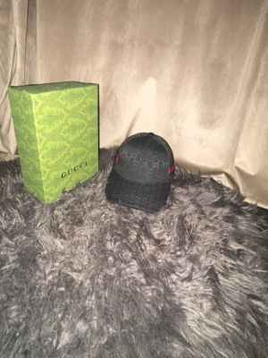 Svart keps från Gucci med monogram - Snygg svart keps från Gucci med klassiskt GG-monogram över hela kepsen och diskreta röda/gröna detaljer på sidan. Justerbar rem baktill och insida med Gucci-logga. Perfekt accessoar för en trendig streetstyle-look.